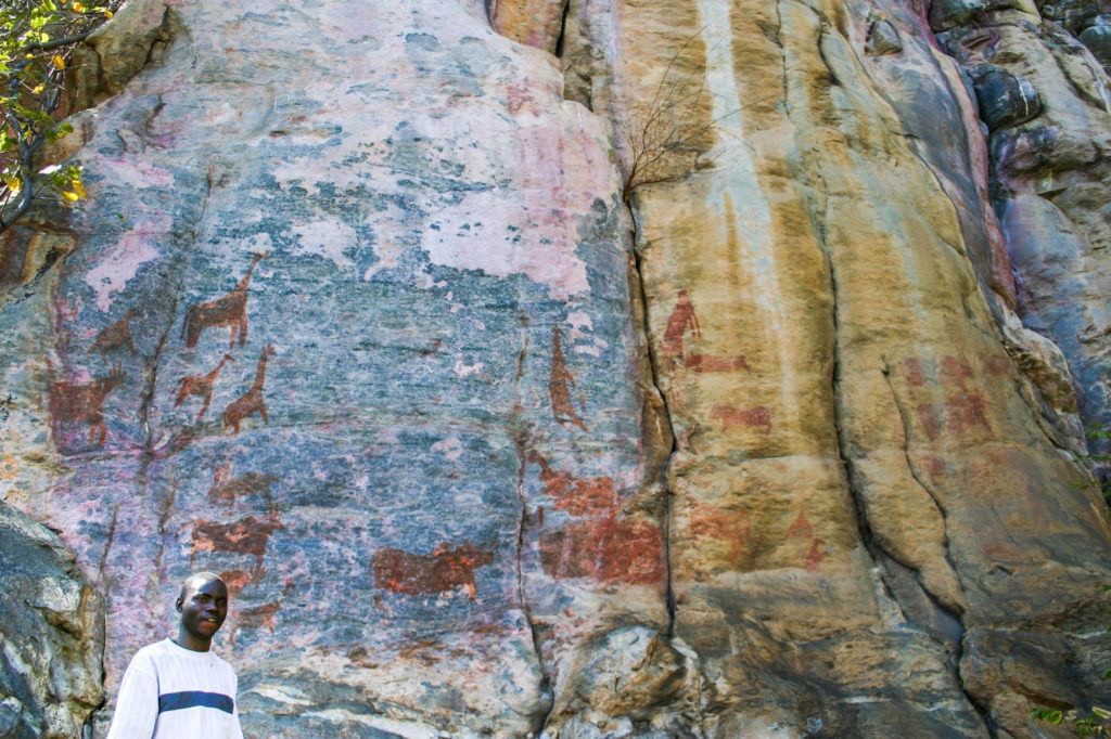 San guide standing below a huge rock with ancient animal figures, including giraffes and antelope at Tsodilo Hills, Botswana.