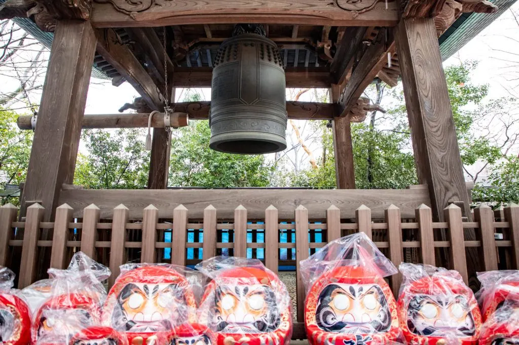 Darumas for sale in front of temple bell.